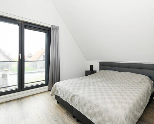 helles schlafzimmer mit großem fenster schwarzen ränder und holzboden sowie weiße wände graue vorhänge und ein bett