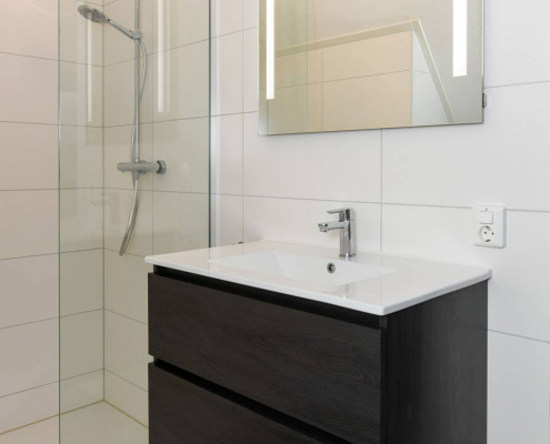 weißes badezimmer mit offener dusche und hellen wandfliesen sowie dunklen bodenfließen und scharzen holzschubladen unter dem waschbecken